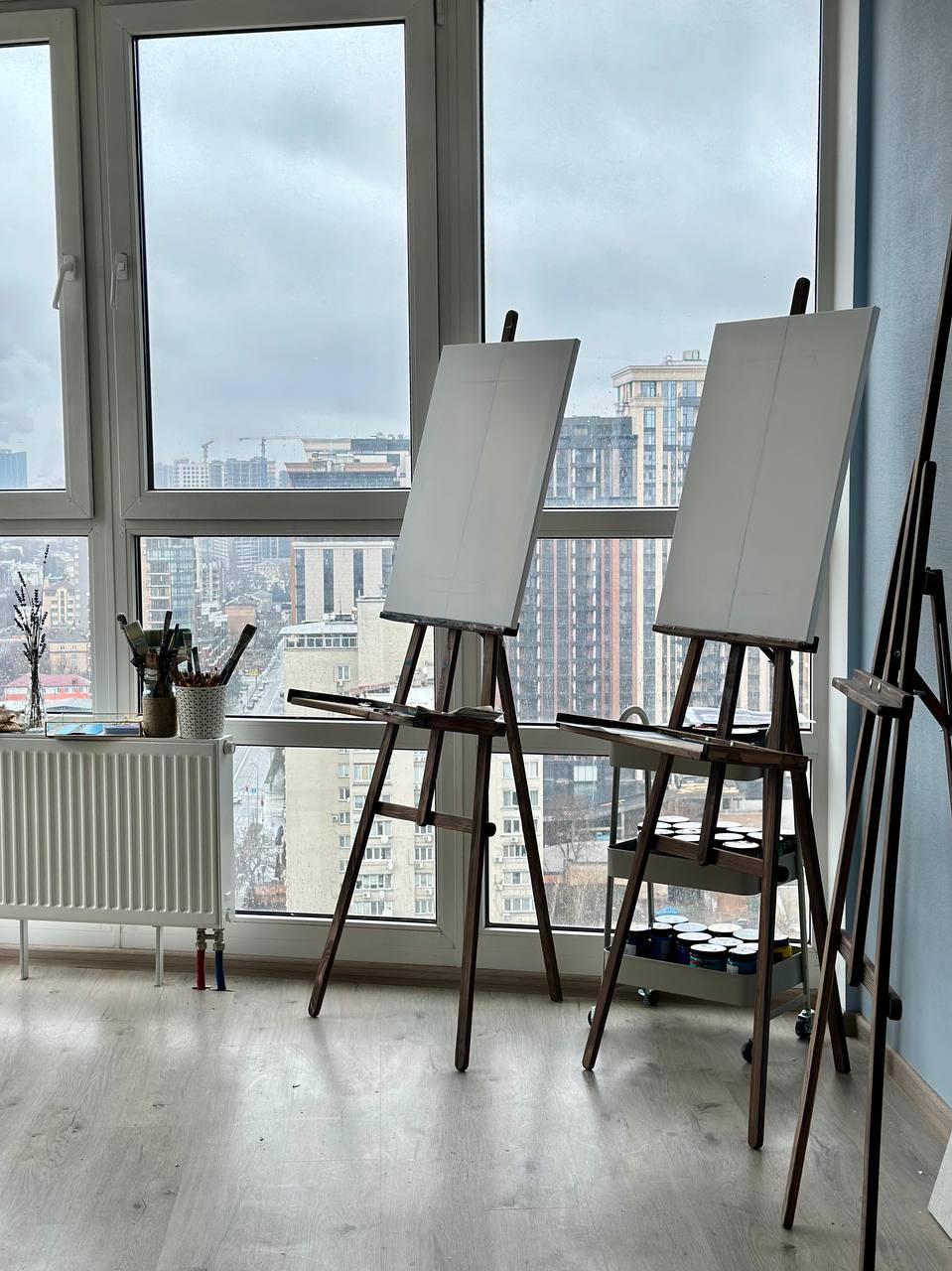 Art easels with blank canvases positioned by large windows overlooking Kyiv cityscape